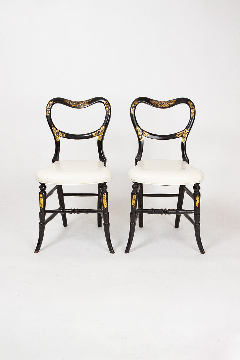 A pair of antique balloon back chairs with white upholstered seats and ornate mother of pearl inlay on ebonized wood frames with gilt details and cabriole front legs and sabre back legs.